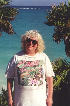 Deborah between palm trees with the sea behind her.